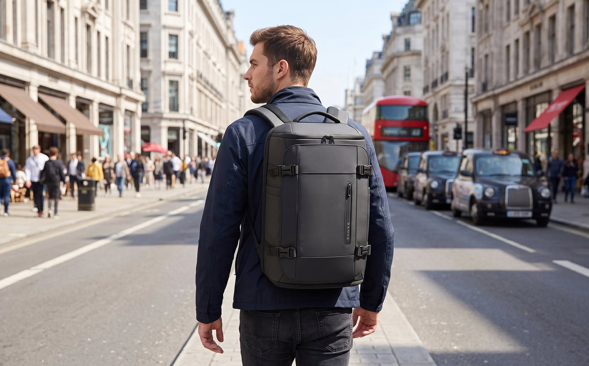 A man wearing a dark grey BANGE backpack walks down a busy city street with London-style buildings, pedestrians, and red double-decker buses in the background.