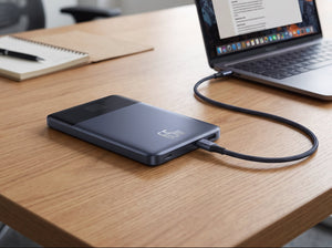 A portable external hard drive in a dark slate blue color is connected to a laptop on a wooden desk in a bright office setting.
