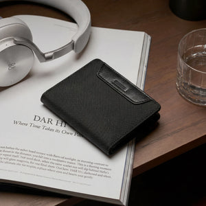 Black slim bifold wallet on open book, textured weave and metal logo tab.