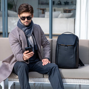 Dark backpack with front flap pocket worn by man seated, showing top handle
