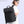 Black travel backpack worn by a person, featuring top flap, front pocket, and shoulder straps.